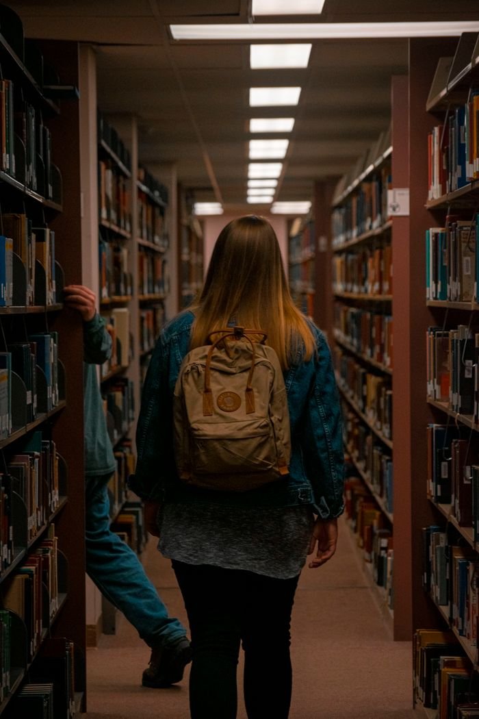 services-06 A young woman with a backpack navigates through bookshelves in a library corridor.