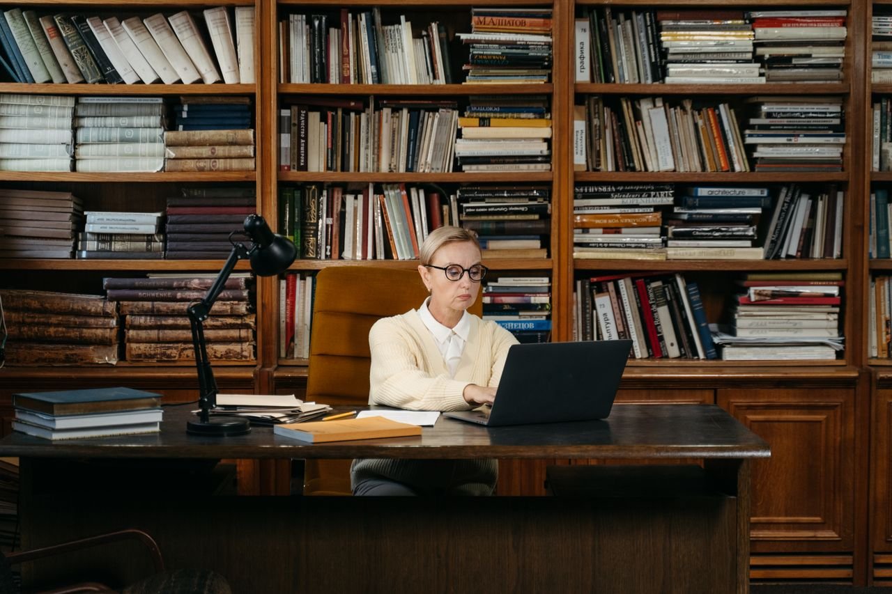 services-05 Woman in a yellow sweater working on a laptop amidst books in a library setting.